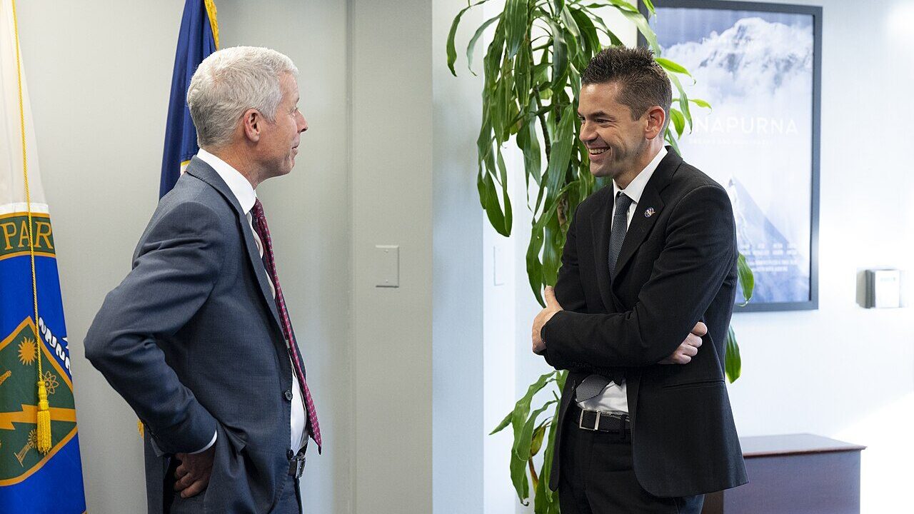 Two officials in suits talking beside national flags inside a government office